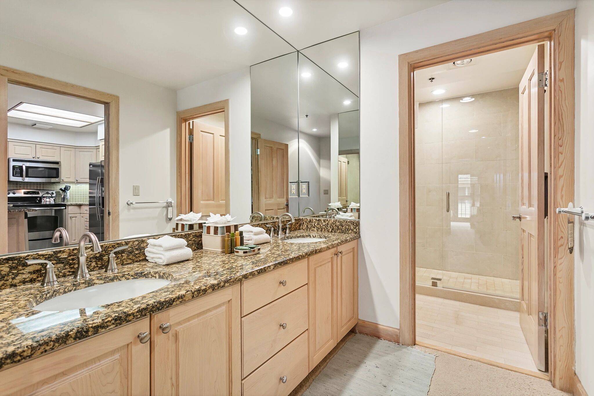 A modern bathroom with a granite countertop, two sinks, a large mirror, and a walk-in shower.