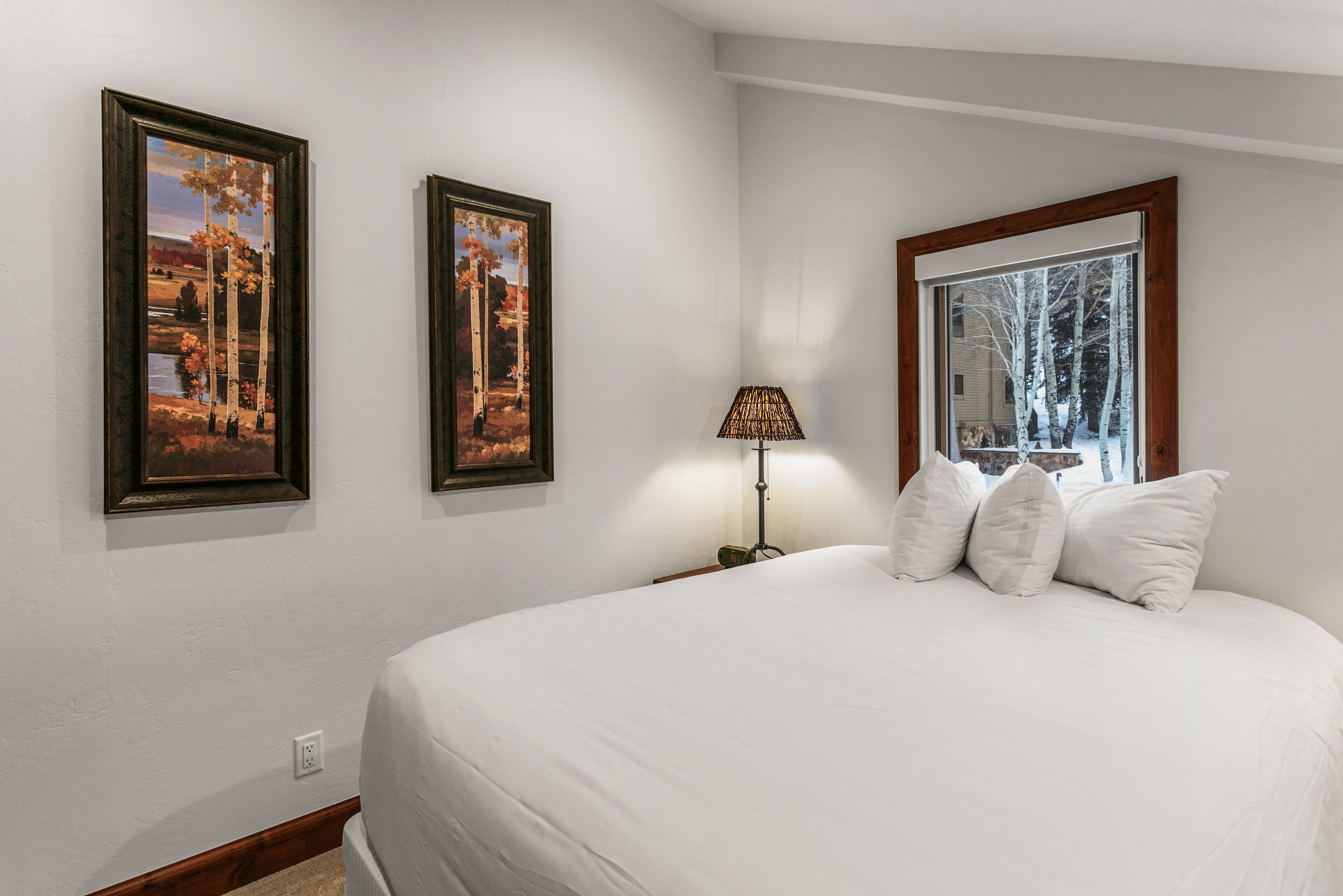 Une chambre à coucher avec un grand lit, deux tableaux encadrés, une lampe de chevet et une fenêtre donnant sur des arbres.