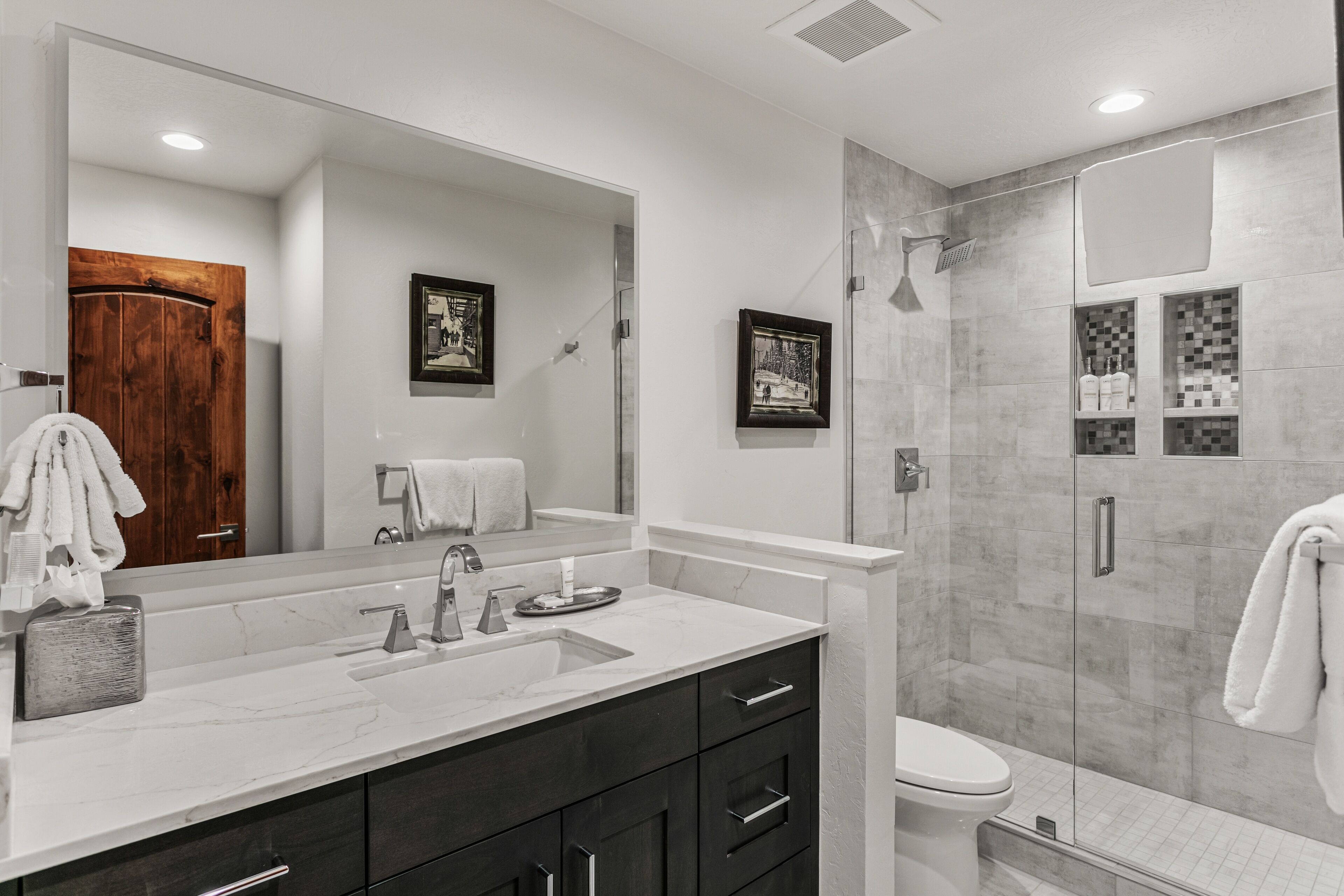 A modern bathroom with a large mirror, a double sink vanity, a toilet, and a shower area.