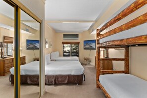 Ein Hotelzimmer mit zwei Betten, einem Schlafplatz in Etagenbetten, einem großen Spiegel, einem Schreibtisch mit Lampe sowie einem an der Wand montierten Fernseher.