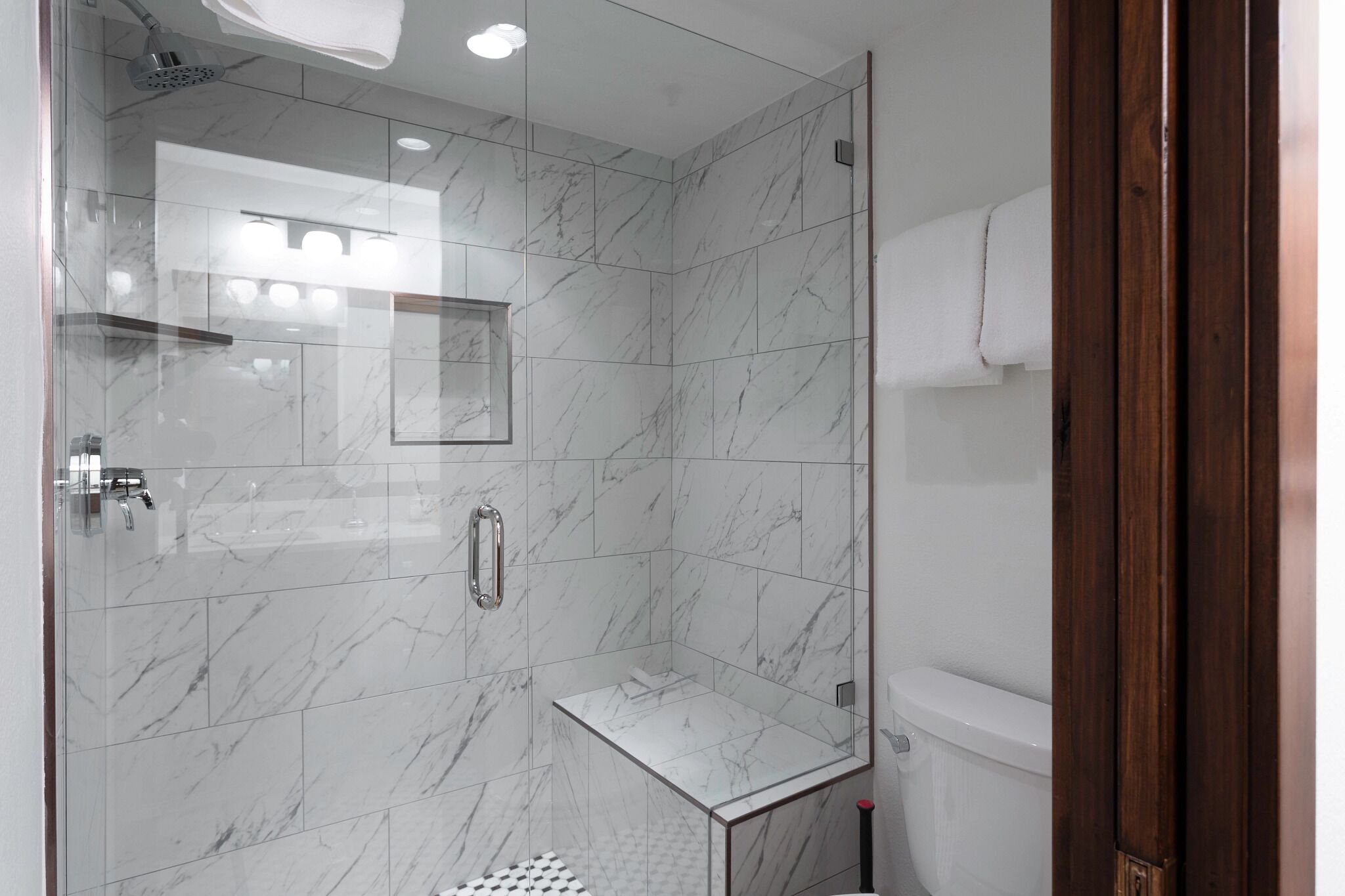 A modern bathroom with a glass-enclosed shower, marble tiles, a toilet, and a wooden door.