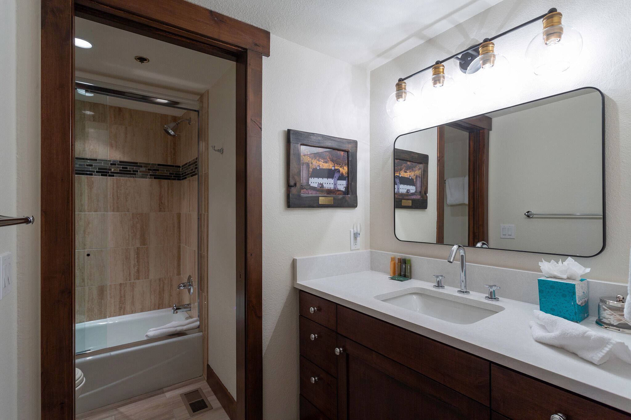 A bathroom with a bathtub, a sink, a vanity with drawers, a large mirror, and a framed picture on the wall.
