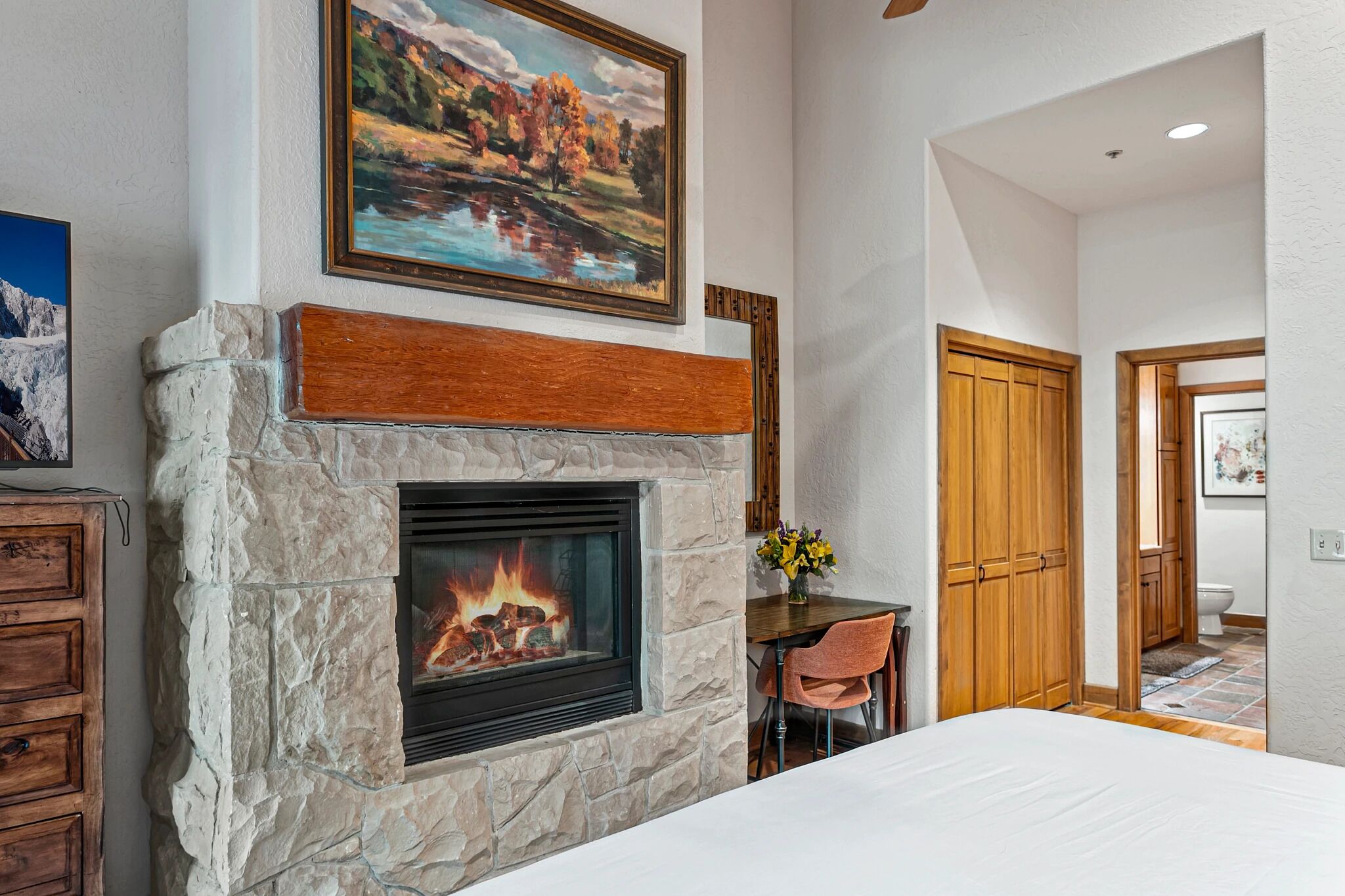 A stone fireplace with a fire, a wooden bench, a dining table with chairs, and a painting above the fireplace.