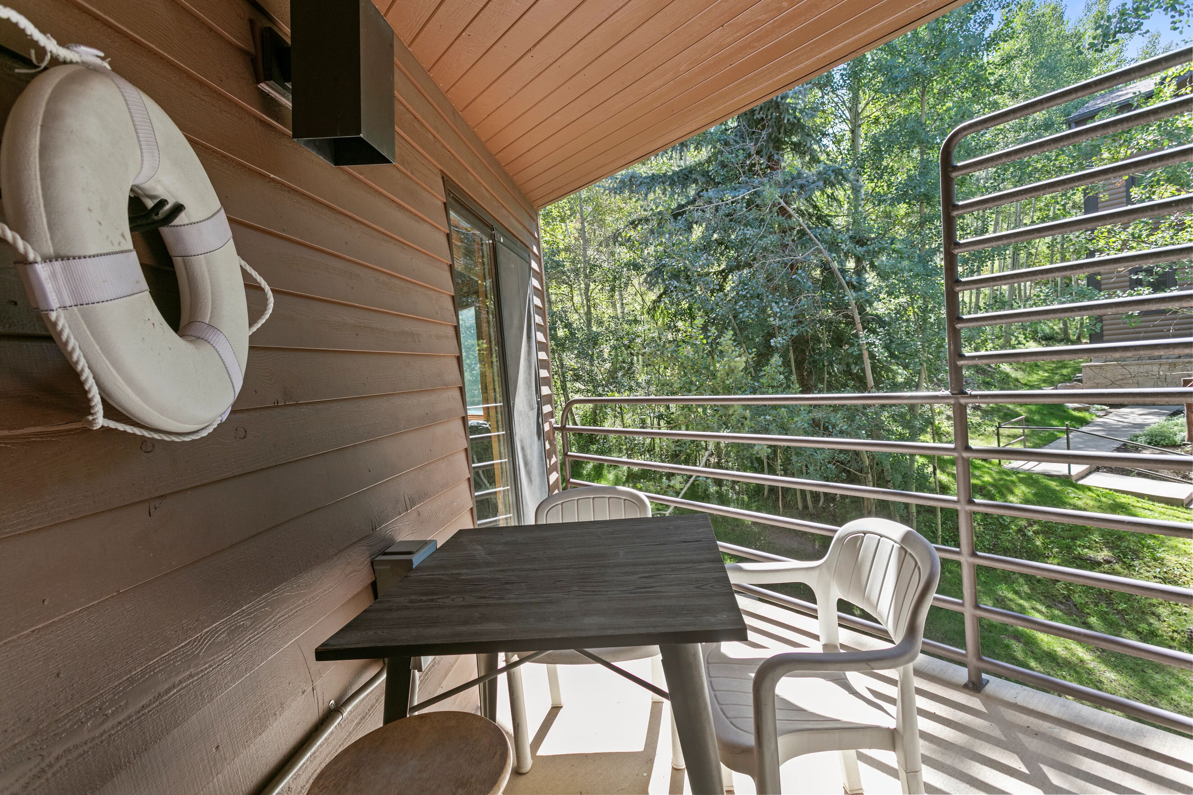 Une terrasse en bois avec une table et des chaises, une bouée de sauvetage accrochée au mur et une vue sur une zone boisée.