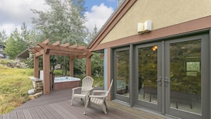 Une terrasse en bois avec des fauteuils Adirondack et un jacuzzi, donnant accès à une entrée par une porte vitrée.