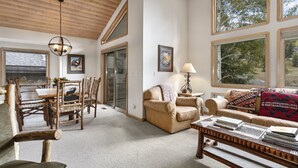 A spacious living area with a high ceiling, wooden beams, a dining table with chairs, a sofa, a coffee table, and large windows offering views of the outdoors. - Boulder Creek at Deer Valley Resort - Three Bedroom Condo with Hot Tub #1119 3 Condo (Park City)