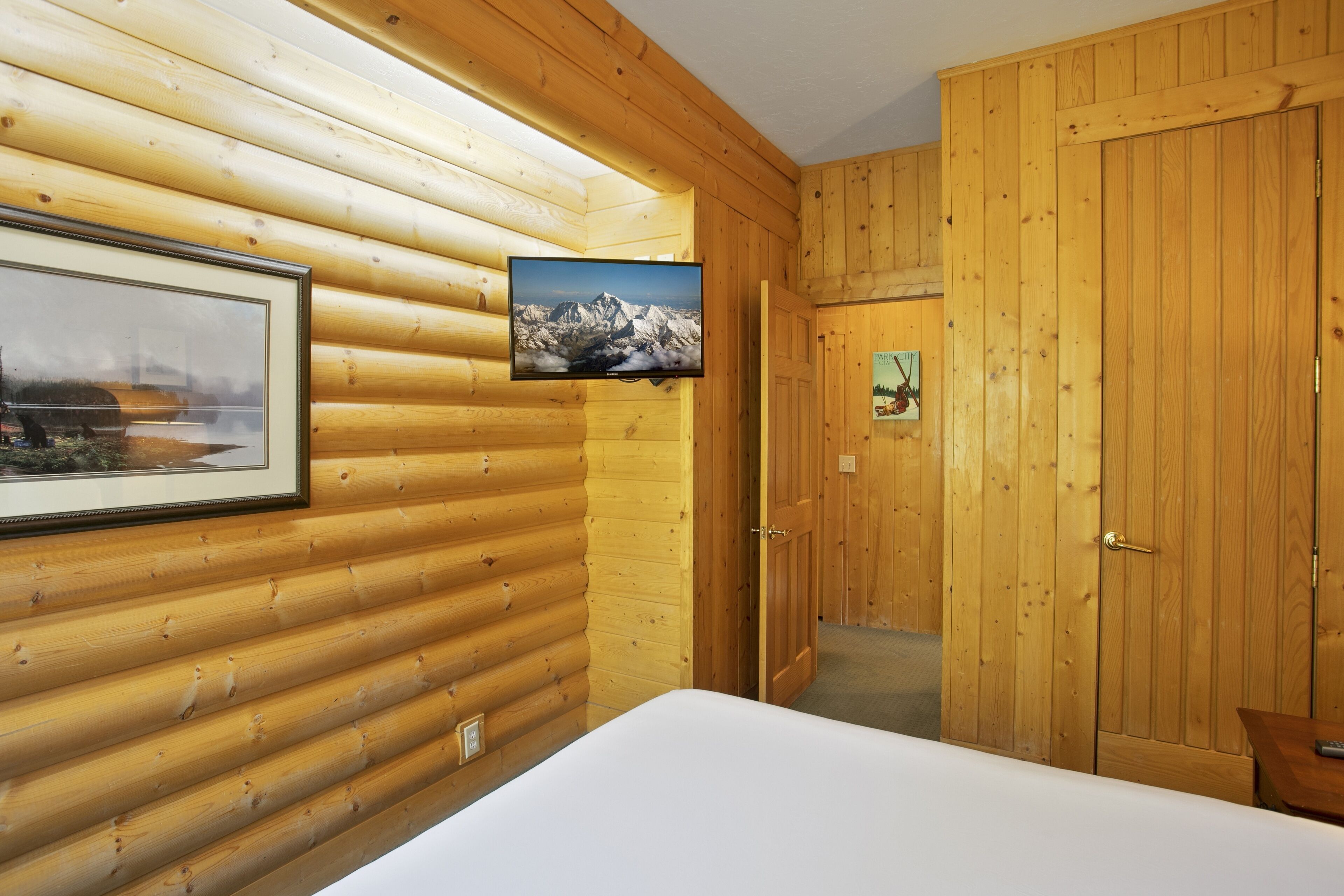 Una camera in una baita di legno con un letto, una TV a parete che mostra una scena montana e un quadro appeso al muro.