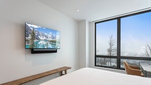 A bedroom with a large window, a bed, a wooden bench, and a TV mounted on the wall displaying a mountain landscape. - Argent at Deer Valley Resort - Three Bedroom Deluxe #405 3 Condo (Park City)
