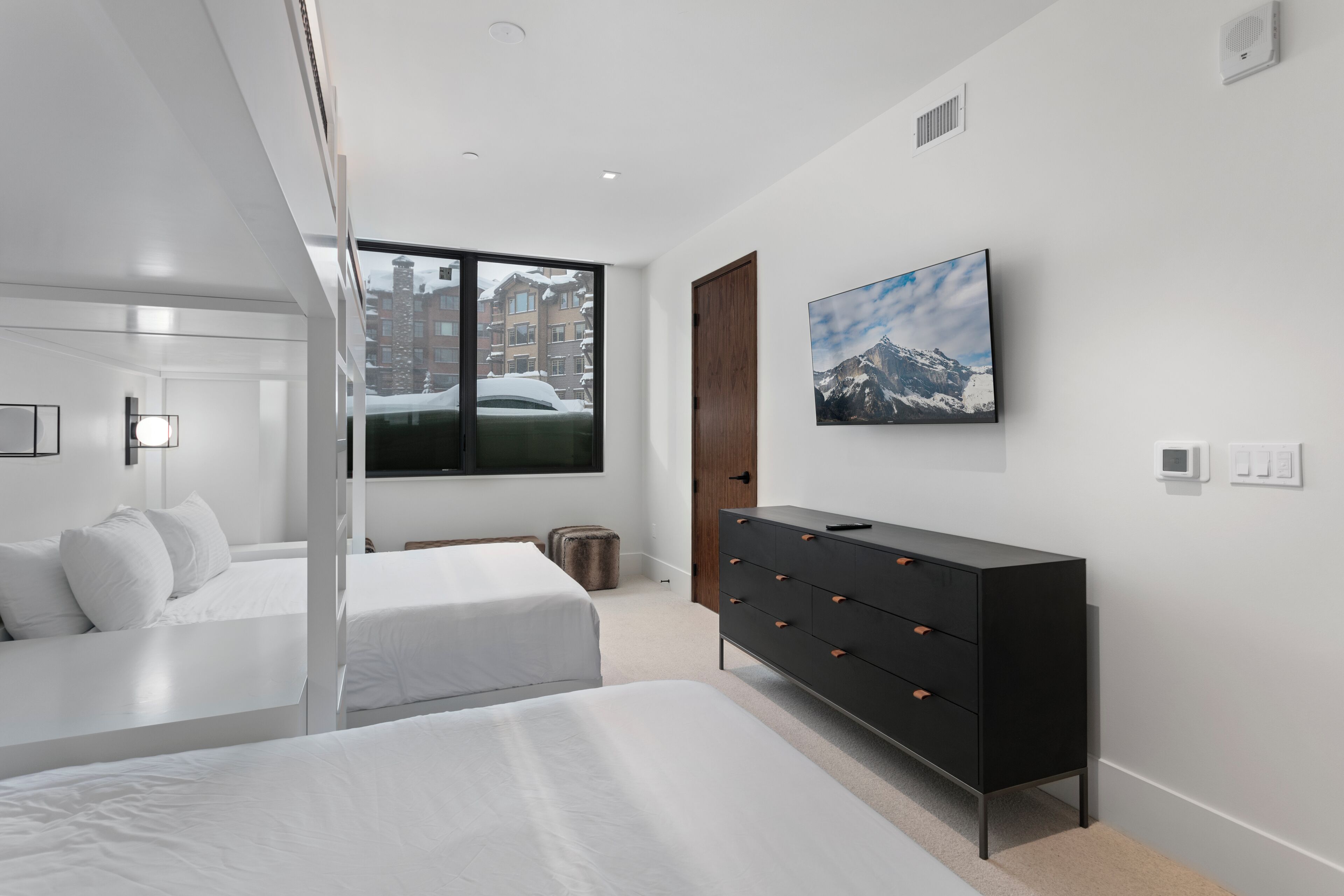 A modern bedroom with two beds, a dresser, a TV, and a window with a view of buildings.