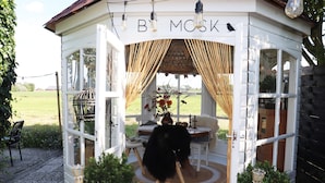 Un bâtiment accueillant, de style cottage, aux murs blancs, sur lequel on peut lire l’inscription « B MCSK ». Il est doté de grandes fenêtres, d’une petite table avec des chaises et d’un vase décoratif rempli de fleurs.