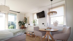 Une chambre moderne et bien éclairée, avec un lit, une table ronde entourée de chaises, un téléviseur fixé au mur et de grandes fenêtres avec des rideaux.