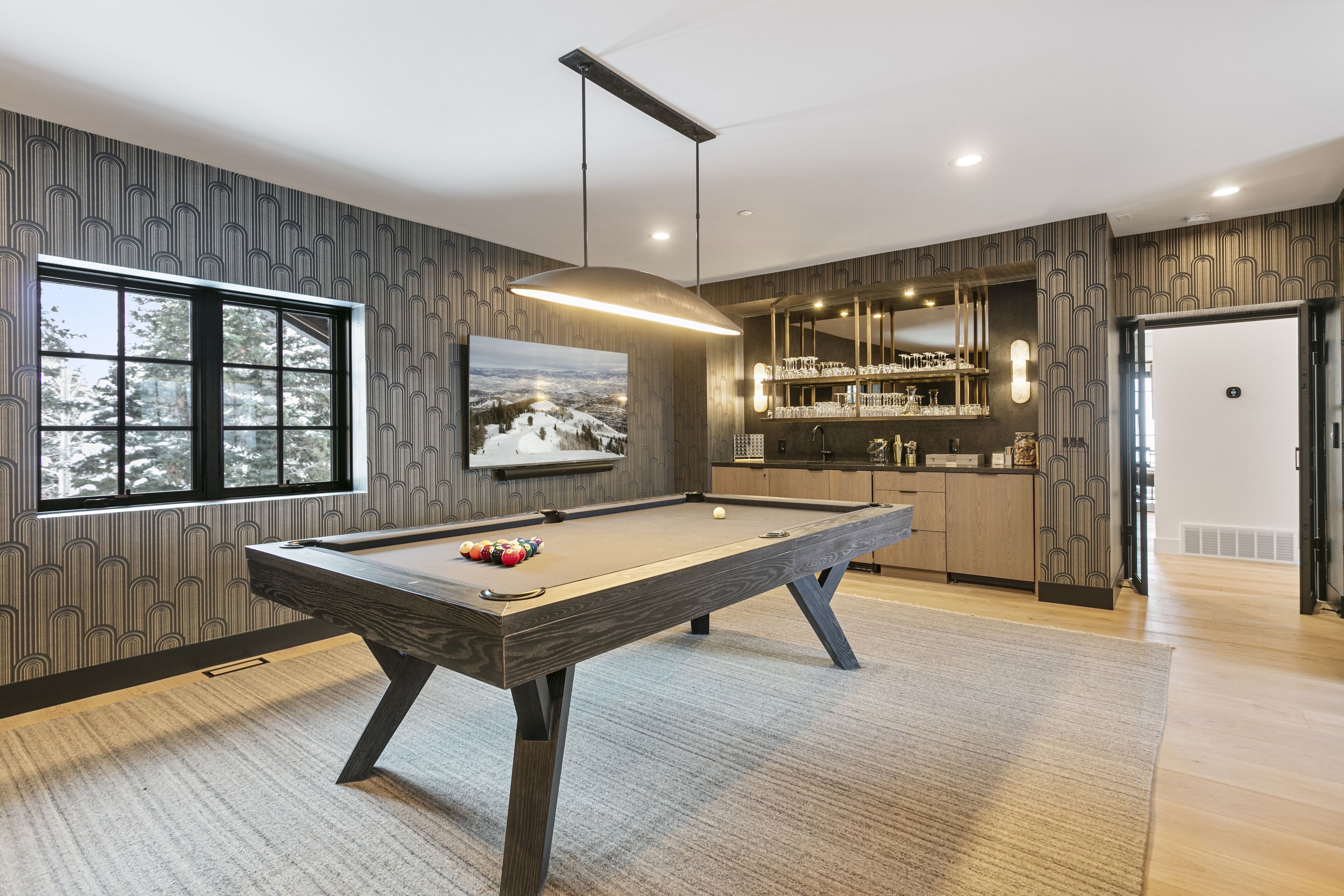 A modern pool table in a well-lit room with a large window, a bar area, and a view of a snowy landscape.
