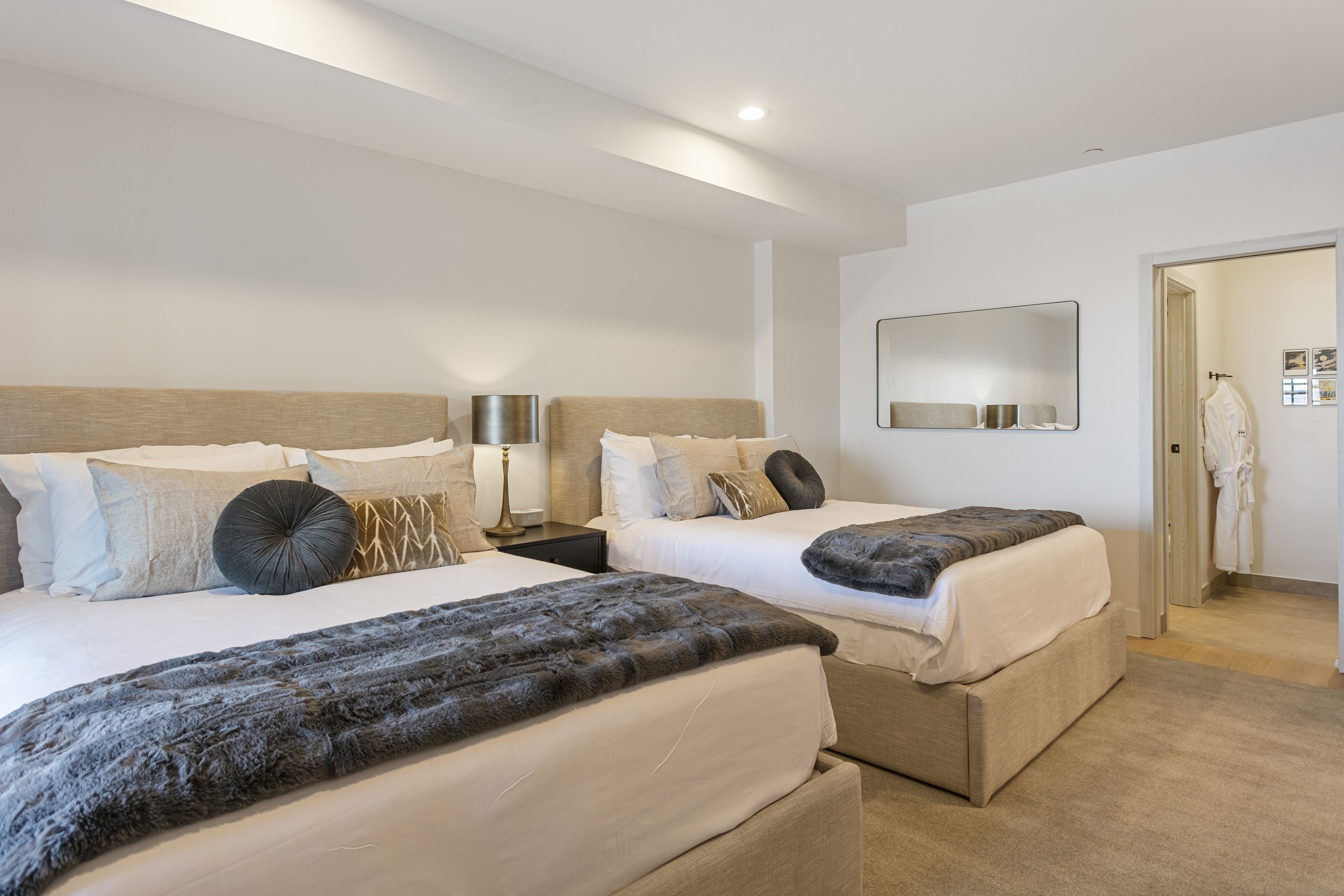 A hotel room with two beds, a bedside table with a lamp, a mirror, and a robe hanging on a door.