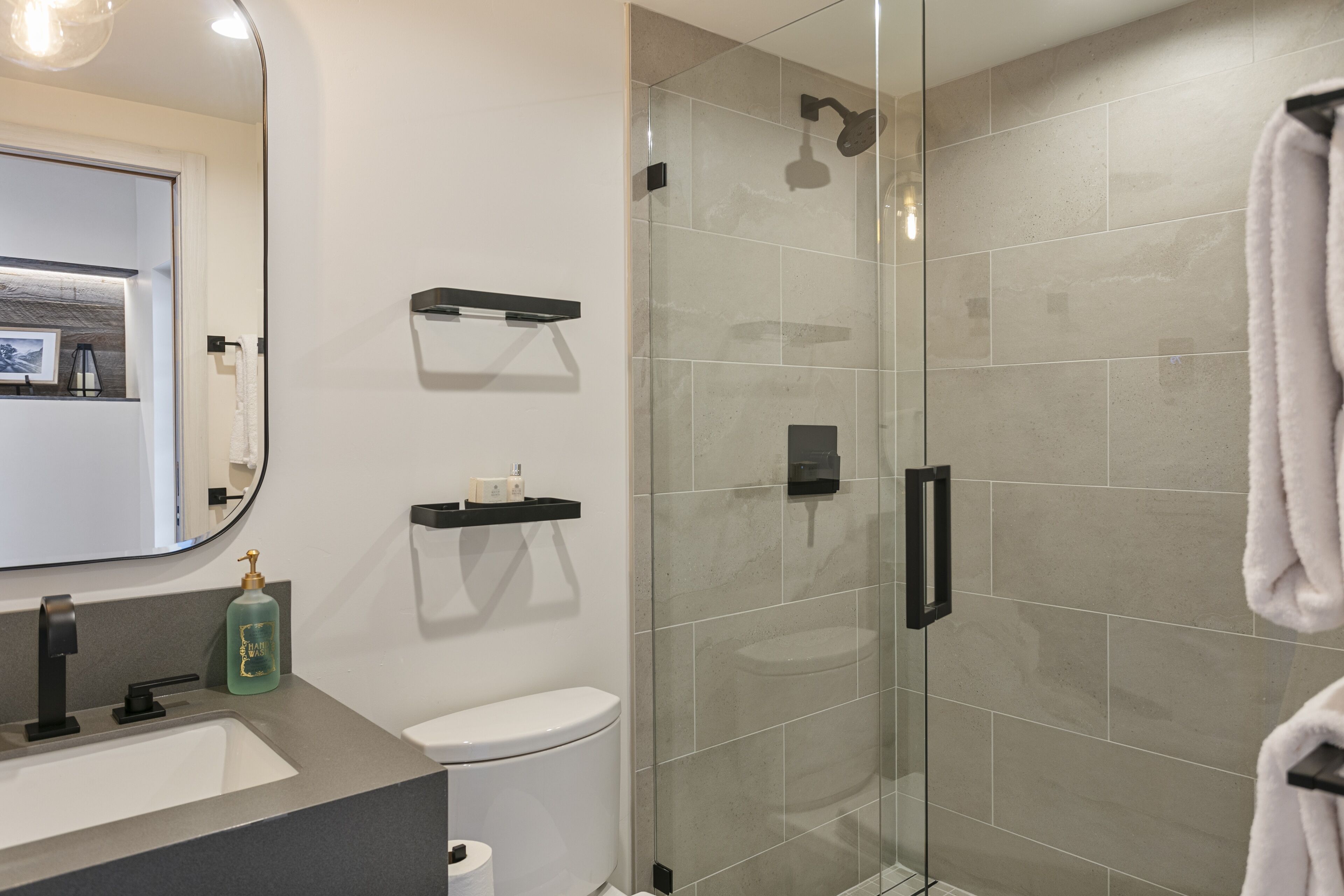 Un baño moderno con ducha de vidrio, inodoro blanco, lavamanos con dispensador de jabón y barra para toallas.