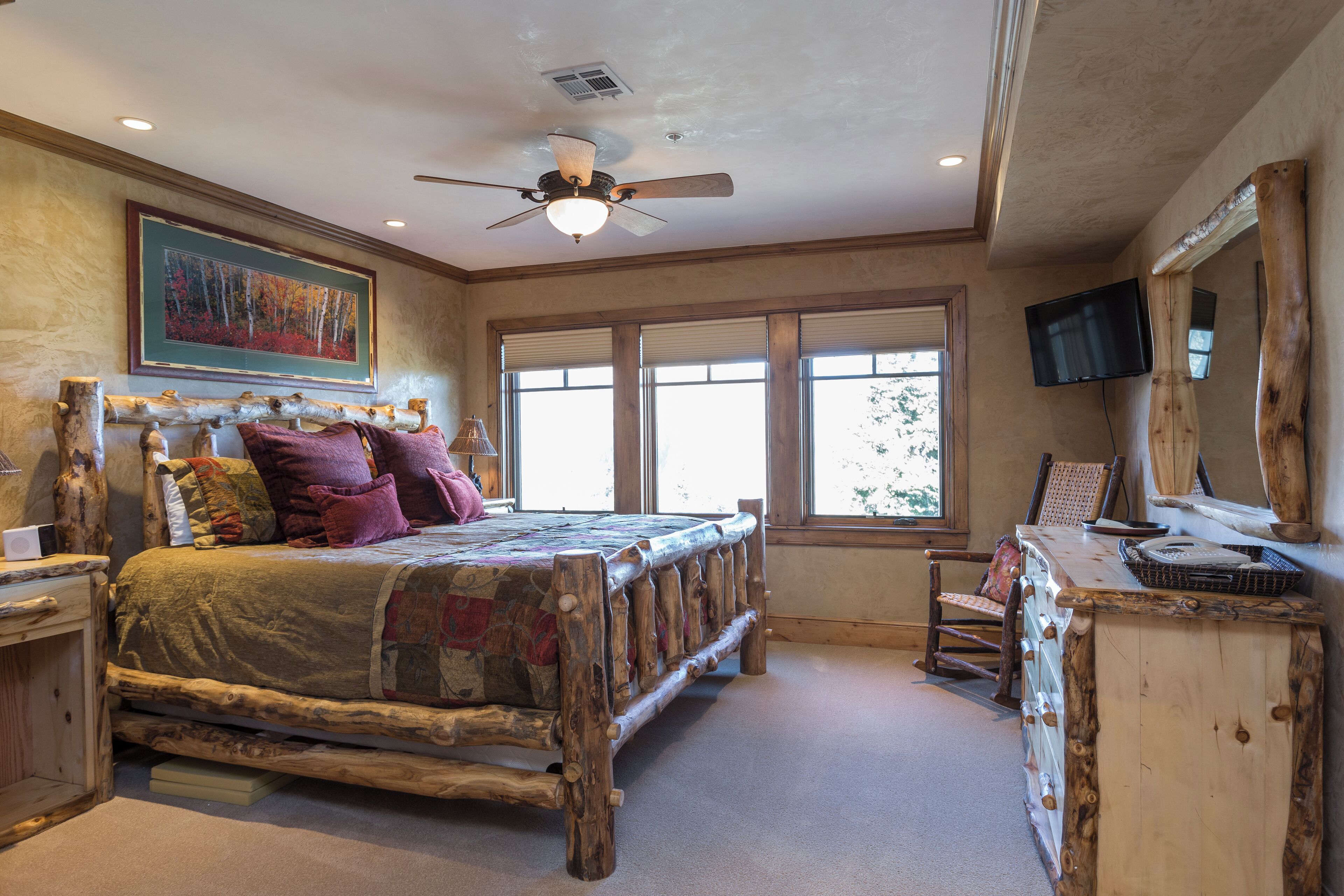 A rustic bedroom with a wooden bed, a ceiling fan, a mounted TV, and large windows.