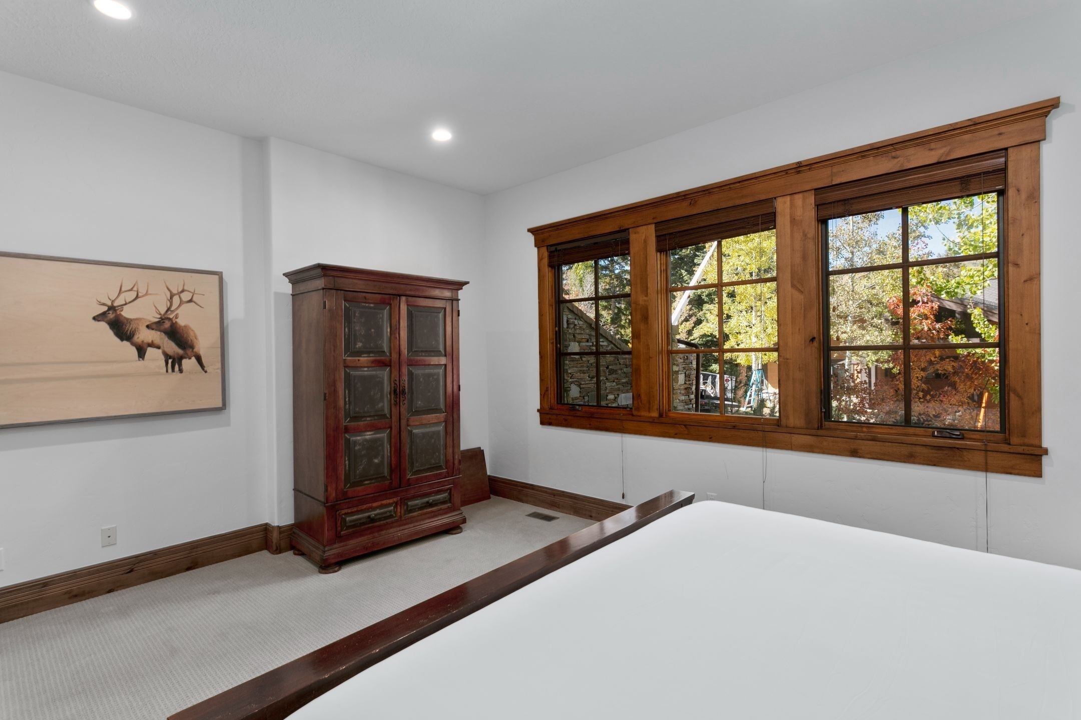 A bedroom with a large window, a wooden cabinet, and a framed deer painting.