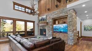 A spacious living room with a stone fireplace, leather sofa, and large windows overlooking a scenic view. - Lookout at Deer Valley Resort - Four Bedroom Residence with Spa #6559 4 Condo (Park City)