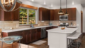 A modern kitchen with dark wooden cabinets, a white island with bar stools, stainless steel appliances, and a large window offering a view of greenery.
