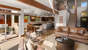 A spacious living area with a leather sofa, a coffee table, and a dining area with chairs and a table. A kitchen with a large island and bar stools is visible in the background.