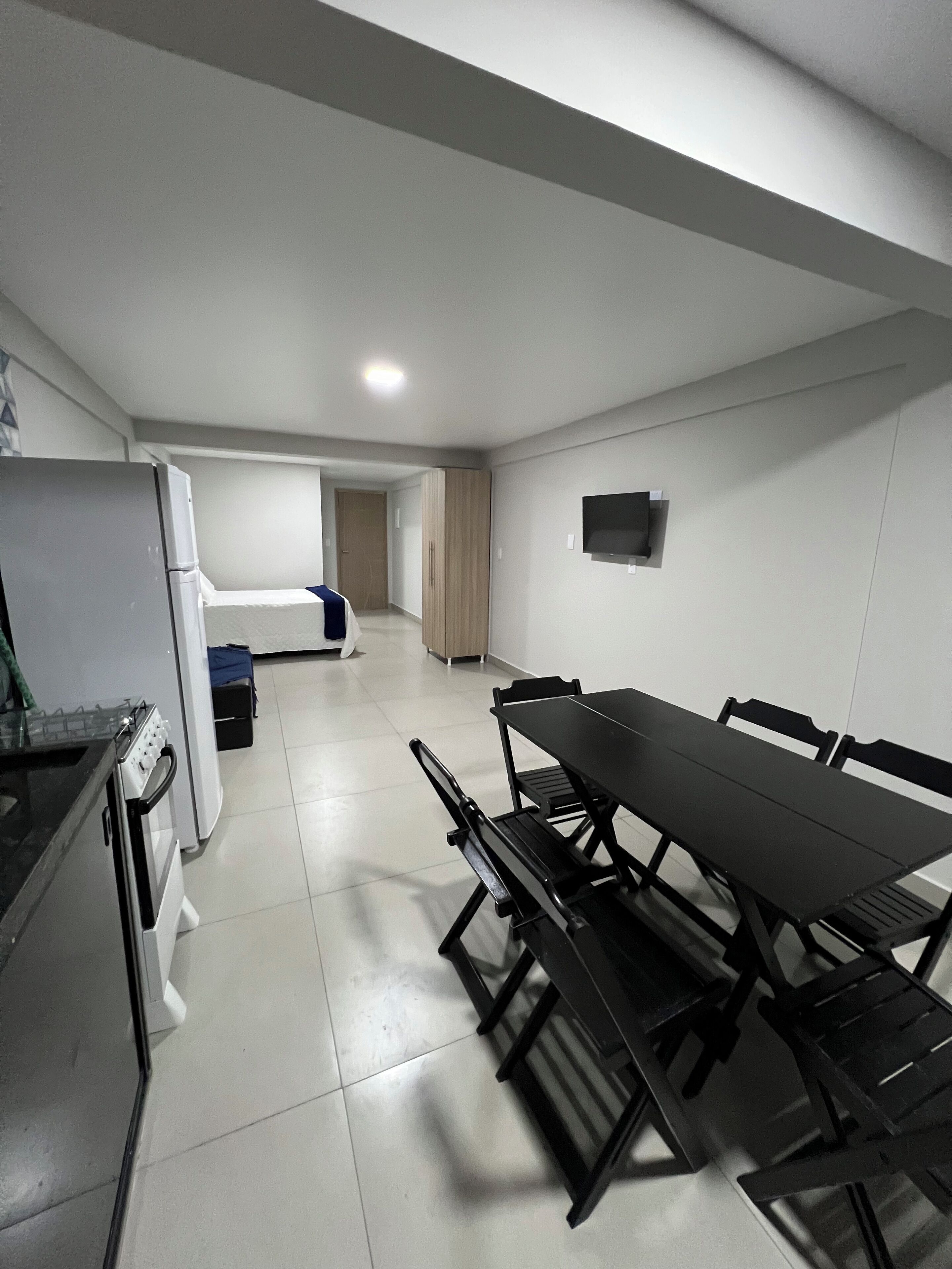 Cocina compacta con refrigerador, estufa y microondas. En primer plano, una mesa de comedor con sillas. Se divisan una cama y un armario al fondo.