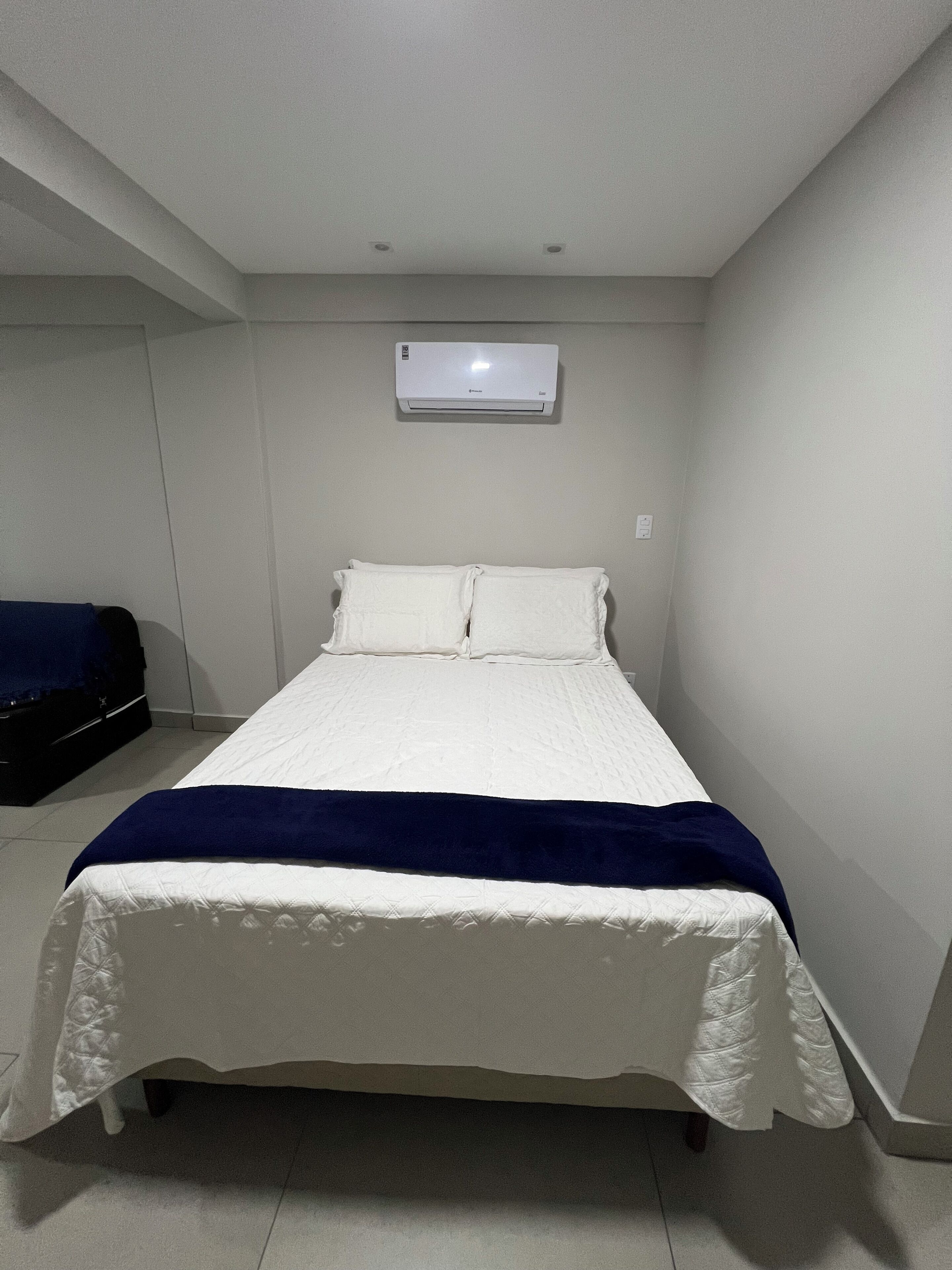 A single-bed room with a white bedspread, a blue blanket, and an air conditioning unit on the wall.