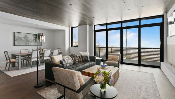 A modern living room with a dark leather sofa, a dining table with chairs, a coffee table with a vase, and large windows offering a view of the outdoors.