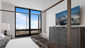 A bedroom with a large window, a fur throw, a bed, a dresser, and a mountain view painting.