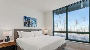 A modern hotel room with a large bed, a nightstand with a lamp, a painting on the wall, and a large window with a view of trees and snow. - Argent At Deer Valley Resort - Hotel Room #105a Hotel Room (Park City)