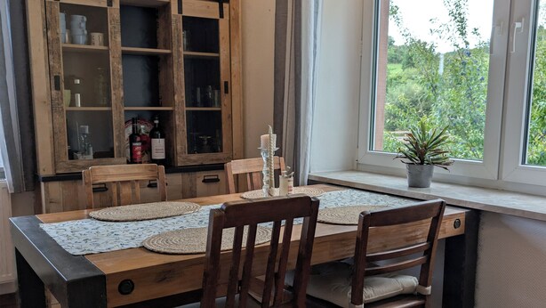 A dining area with a wooden table and chairs, a cabinet with glass doors, and a window with a view of greenery. - Vacation apartment in Bungert - directly on the Eifelsteig trail (Üxheim)