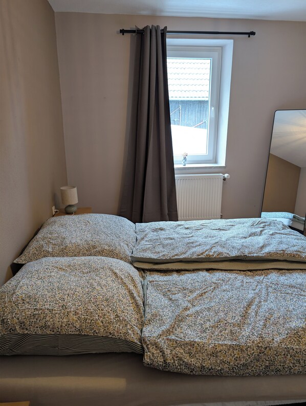A bedroom with a bed, a window with curtains, a radiator, and a bedside table with a lamp. - Vacation apartment in Bungert - directly on the Eifelsteig trail (Üxheim)