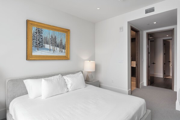 A hotel room with a bed, a nightstand with a lamp, a framed painting of a snowy forest, and a hallway leading to another room.