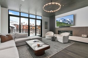 A modern living room with a sectional sofa, a coffee table, and two armchairs. A large flat-screen TV is mounted on the wall above a fireplace.