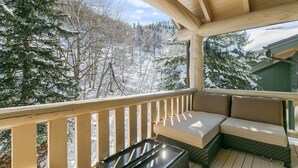 Une terrasse en bois avec un coin salon doté de coussins et une table en verre, offrant une vue sur un paysage enneigé et des arbres.
