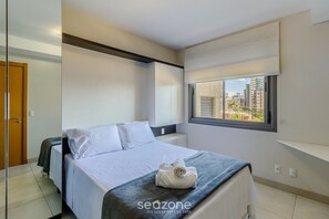A modern bedroom with a bed, a window with a city view, and a built-in wardrobe. - Apto Moderno c/ Churrasq. e Garagem UCR0505 (Porto Alegre)