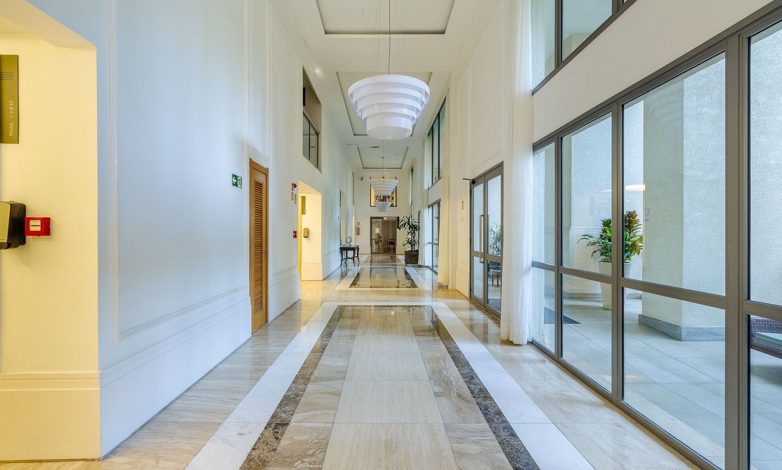 A hotel corridor with a marble floor, large windows, and a modern light fixture.