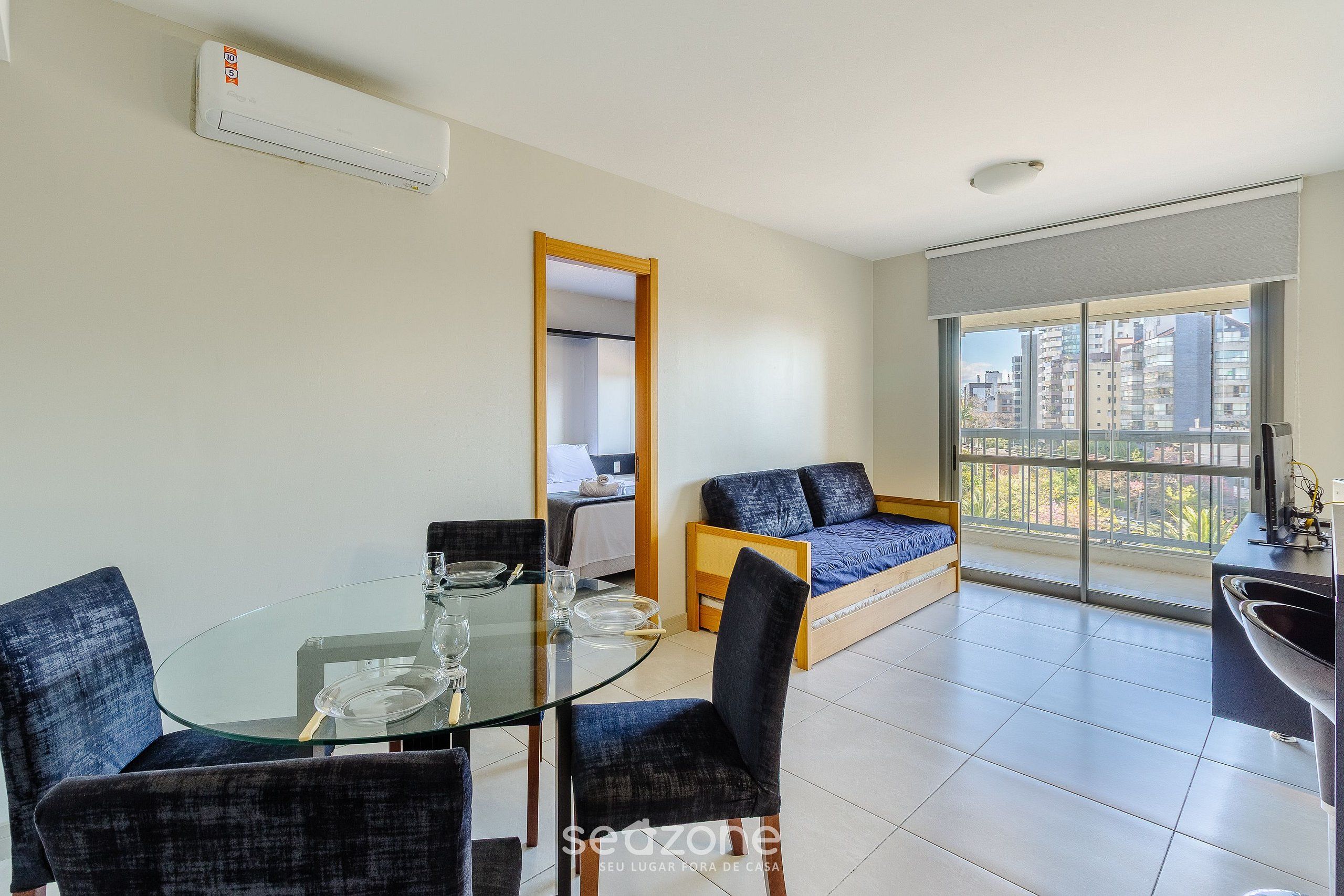 Una sala moderna con una mesa de comedor de vidrio, sillas tapizadas en azul, un sofá y un ventanal con vistas a la ciudad.