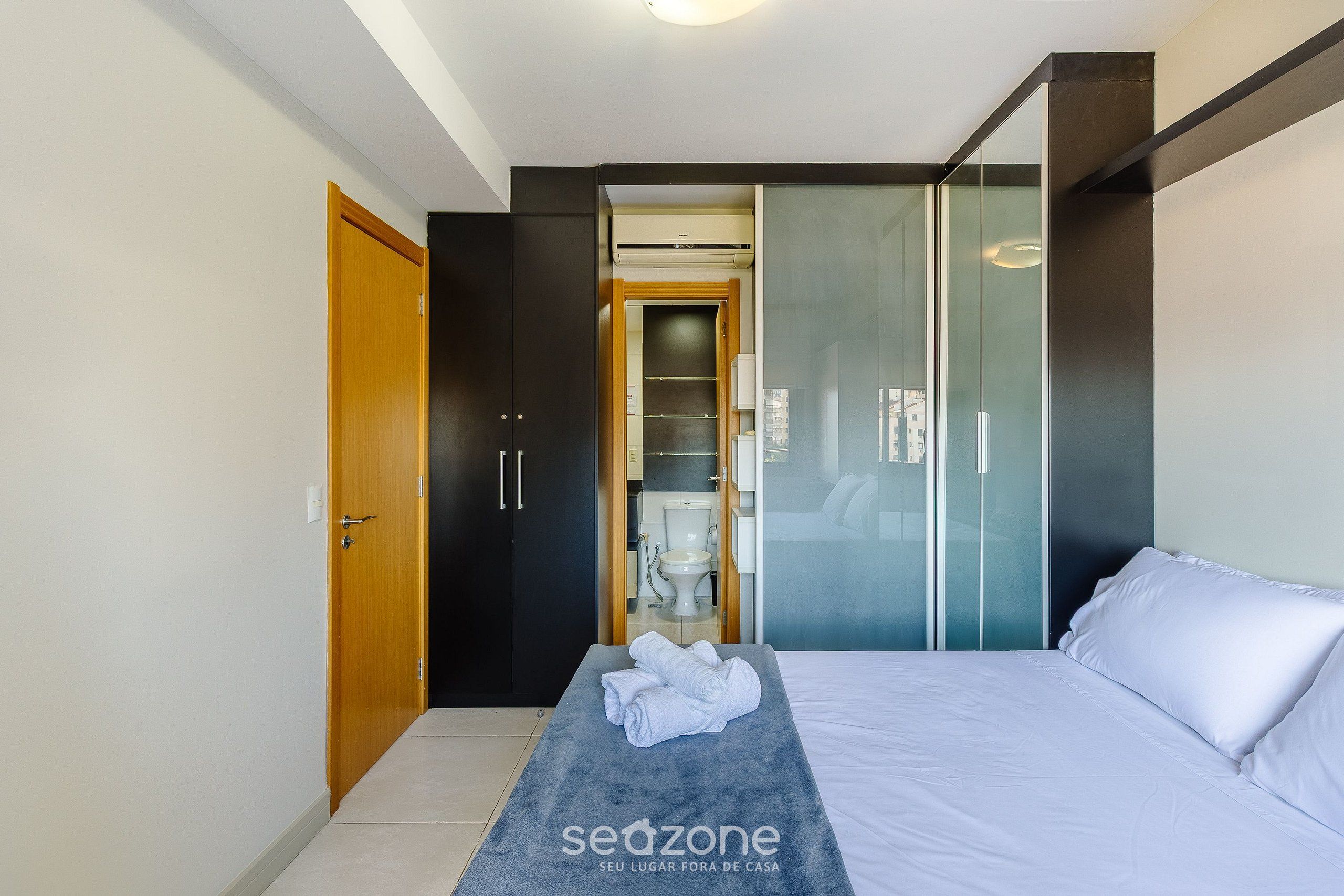 Quarto de hotel moderno com cama, um banco com toalhas, guarda-roupa e divisória de vidro.
