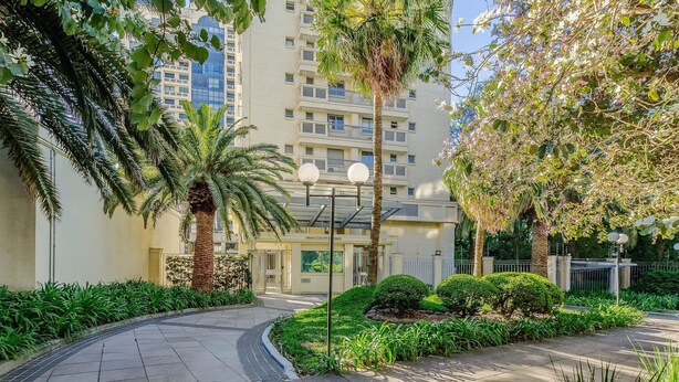 A residential building with a paved walkway, palm trees, and well-maintained greenery. - Apto Moderno c/ Churrasq. e Garagem UCR0505 (Porto Alegre)