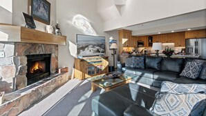 Ein modernes Wohnzimmer mit Kamin aus Stein, einem Glastisch und einem Ledersofa mit gemusterten Kissen.
