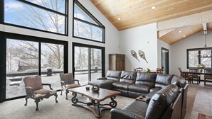 A spacious living room with a large sectional sofa, a wooden coffee table, and two armchairs. Large windows provide a view of the outdoors.