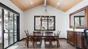 A dining area with a wooden table, chairs, a chandelier, a large window with a view of snow-covered trees, and a wooden cabinet with a mirror.