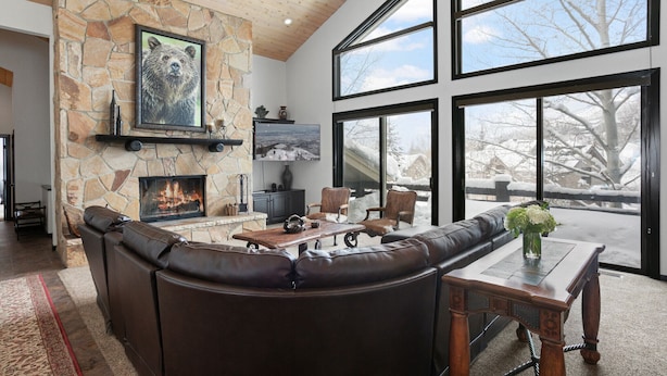 A cozy living room with a stone fireplace, leather sofa, and large windows offering a view of a snowy landscape.