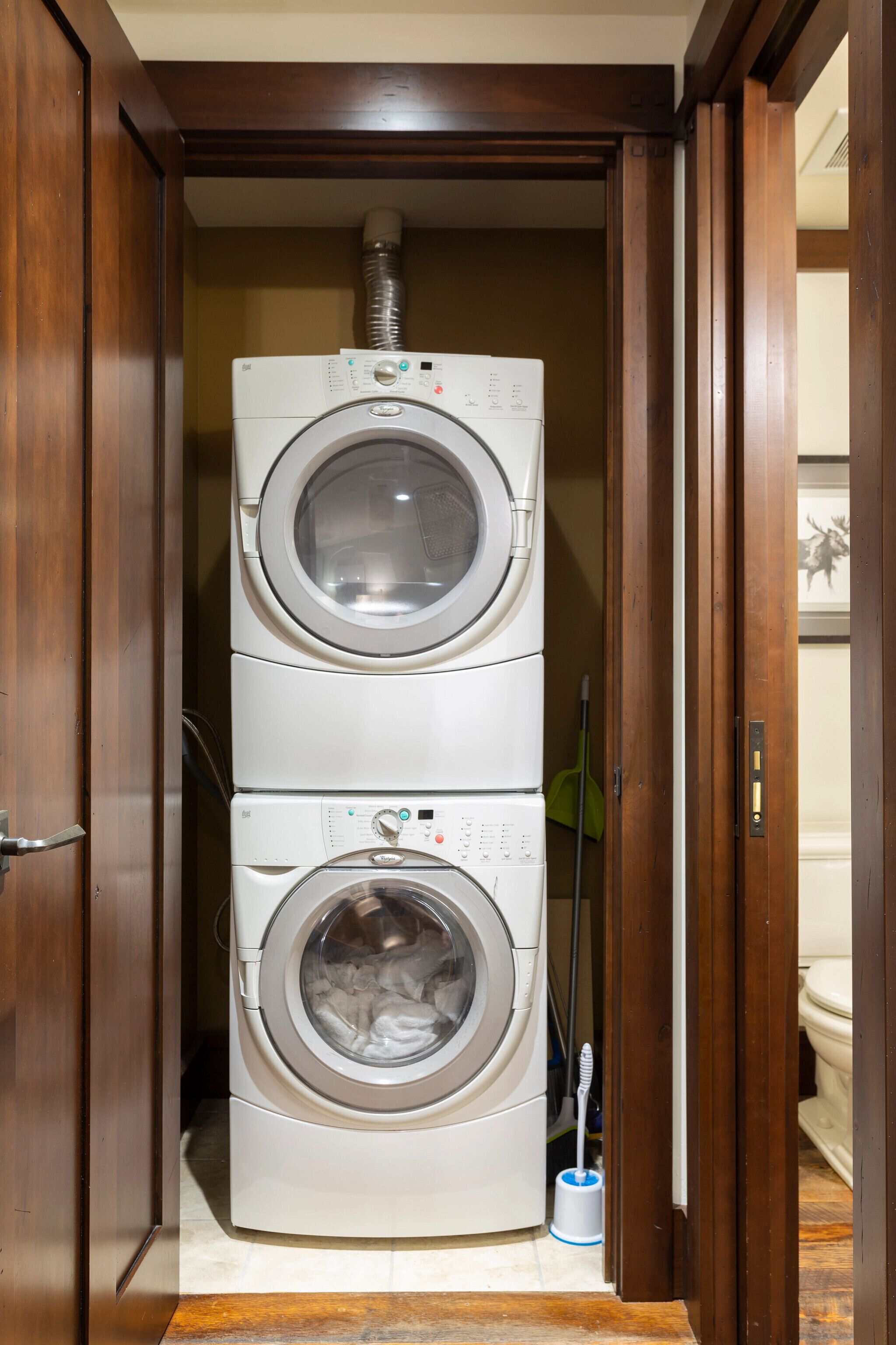A laundry room with stacked washers and dryers, a broom, and a toilet visible through an open door.