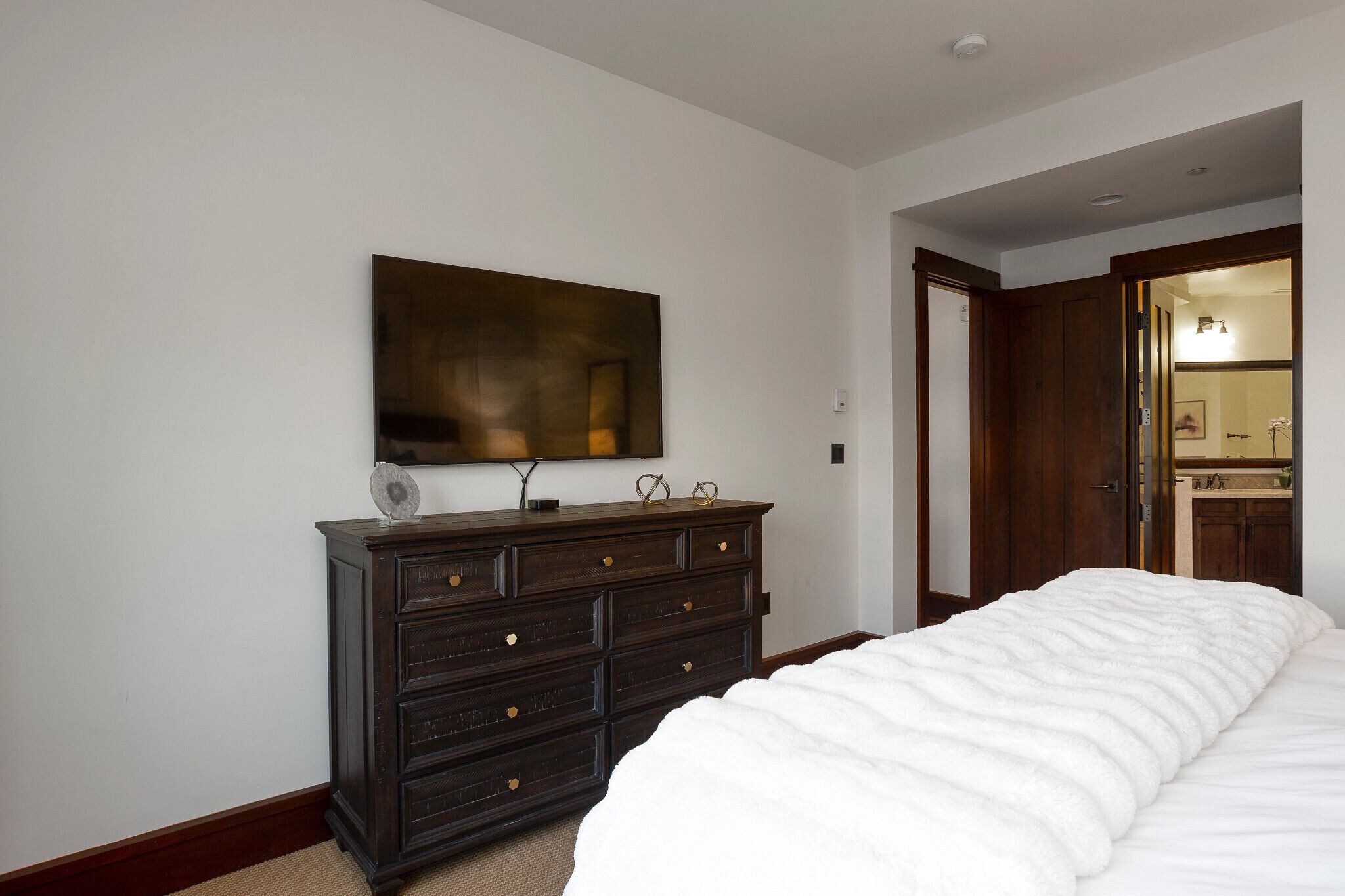 Un dormitorio con una cama grande, un aparador de madera oscura, un televisor montado en la pared y una puerta que da a un baño.