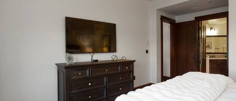 Un dormitorio con una cama grande, un aparador de madera oscura, un televisor montado en la pared y una puerta que da a un baño.