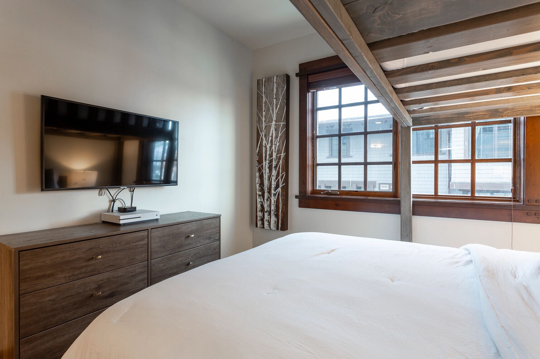 A bedroom with a bed, a dresser, a TV mounted on the wall, and a window with a view of buildings.