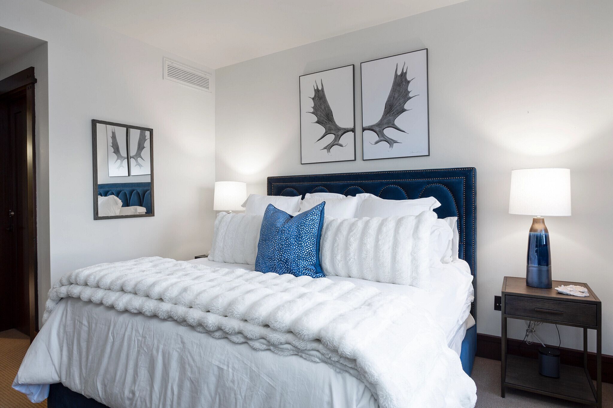 Un lit bien fait, avec une literie blanche et bleue, une tête de lit bleu foncé et deux lampes fixées au mur.