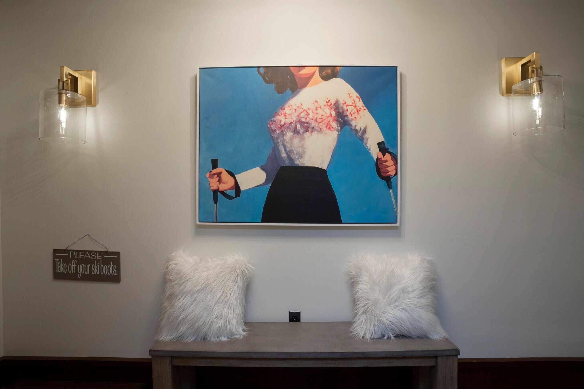 Ein Gemälde zeigt eine Frau mit Skistöcken an einer Wand, ein Schild mit der Aufschrift 'Bitte Skischuhe ausziehen' und zwei weiße, flauschige Kissen auf einem Tisch.