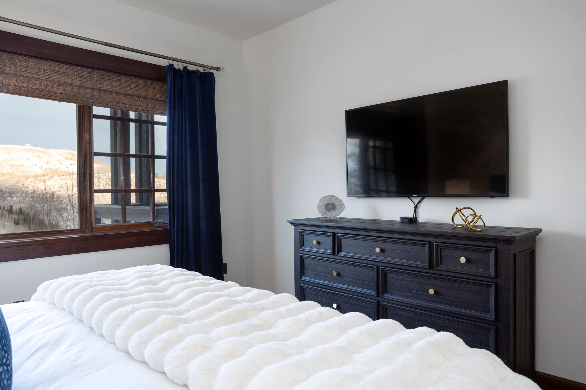 Een slaapkamer met een groot bed, een dressoir, een flatscreen televisie en een raam met uitzicht op een heuvel.