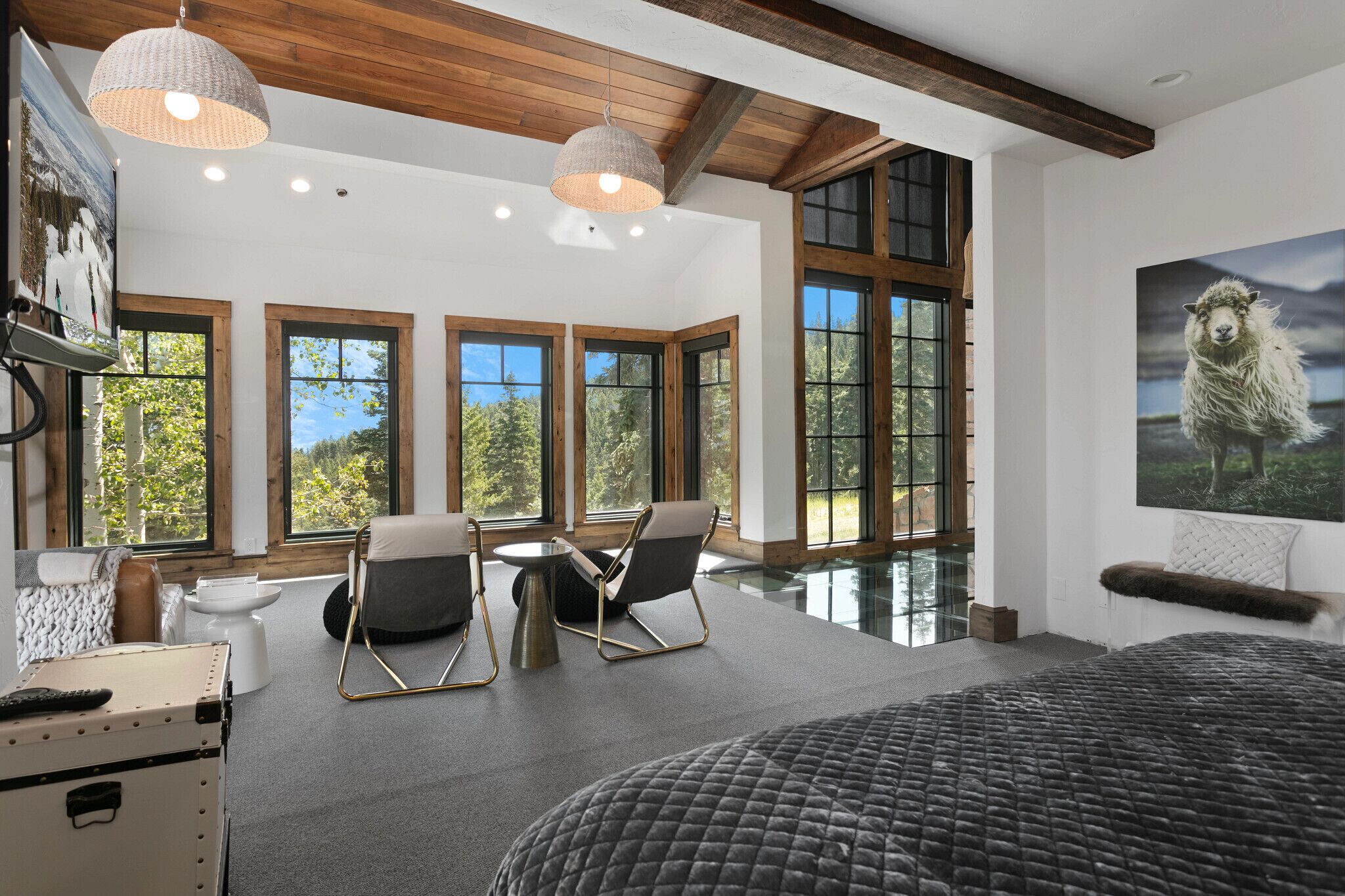 A spacious bedroom with large windows, a bed, a seating area with chairs, and a painting of a sheep.